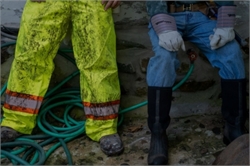 [ai] Two individuals sitting next to each other, one wearing a bright yellow, muddy pair of rain pants with reflective stripes, and the other in blue jeans and black rubber boots. Green hoses are visible in the background.